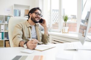 Cheerful communicative young Asian man with beard sitting at table in office and calling customer on phone