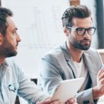 Discussing new business strategy. Young handsome man wearing glasses gesturing and discussing something while other young man holding touchpad and listening to him sitting at the office table