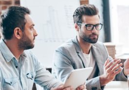 Discussing new business strategy. Young handsome man wearing glasses gesturing and discussing something while other young man holding touchpad and listening to him sitting at the office table