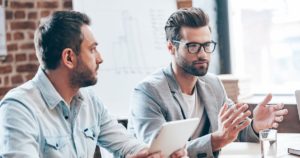 Discussing new business strategy. Young handsome man wearing glasses gesturing and discussing something while other young man holding touchpad and listening to him sitting at the office table