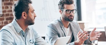 Discussing new business strategy. Young handsome man wearing glasses gesturing and discussing something while other young man holding touchpad and listening to him sitting at the office table