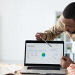 Afro-American marketer pointing at laptop screen while reporting marketing statistics at meeting