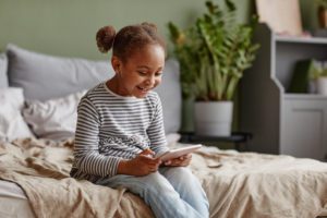 Portrait of cute African-American girl using digital tablet and playing mobile games while sitting on bed, copy space
