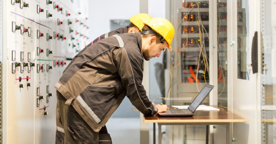 Two maintenance engineers inspect relay protection system with laptop computer. Bay control unit. Medium voltage switchgear