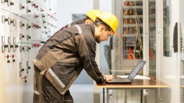 Two maintenance engineers inspect relay protection system with laptop computer. Bay control unit. Medium voltage switchgear