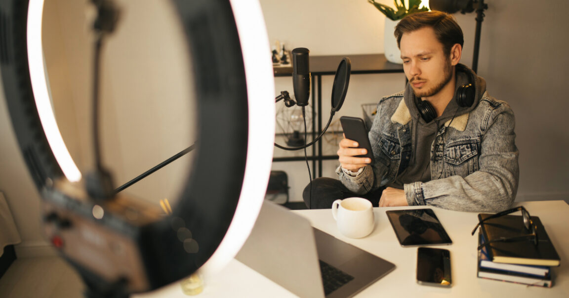 A young man under ring light looking at his phone anxiously with his computer, notebook and iPad on the desk