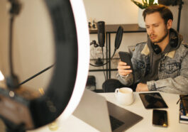 A young man under ring light looking at his phone anxiously with his computer, notebook and iPad on the desk
