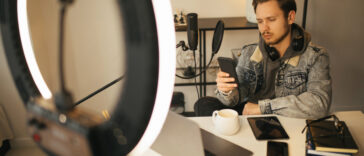 A young man under ring light looking at his phone anxiously with his computer, notebook and iPad on the desk
