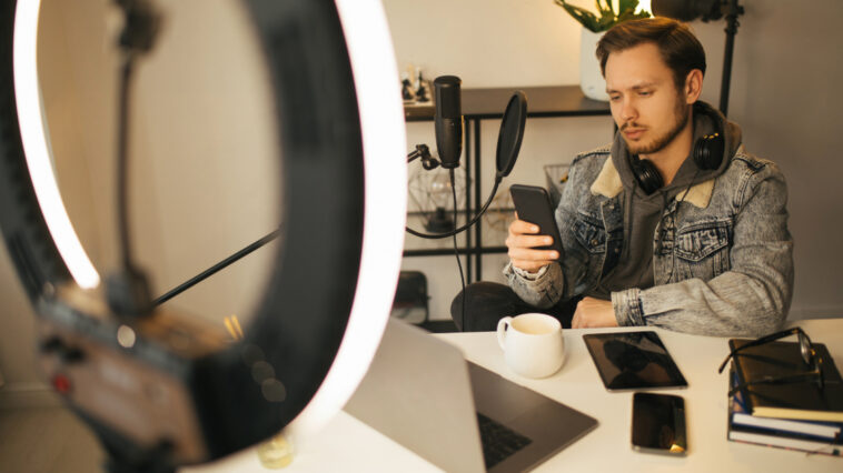 A young man under ring light looking at his phone anxiously with his computer, notebook and iPad on the desk